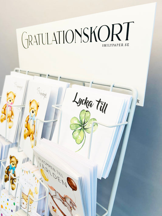 A säsongsställ rack displays smilypaper.se Barnkort greeting cards. The sign reads GRATULATIONSKORT. Designs include teddy bears and a green four-leaf clover card with Lycka till (Good luck) for birthdays.